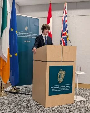 Gabriel Dalton speaking at a podium at the Consulate General of Ireland in Vancouver, beside the Irish, European Union, Canadian and British Columbia flags