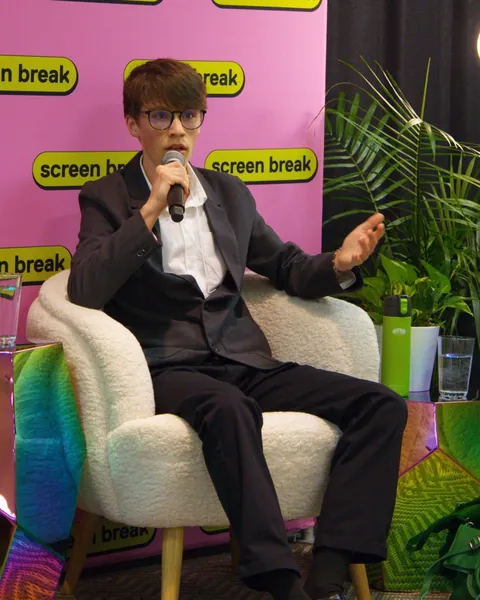 Gabriel Dalton speaking into a microphone on the URL to IRL panel, seated in front of a pink Screen Break backdrop